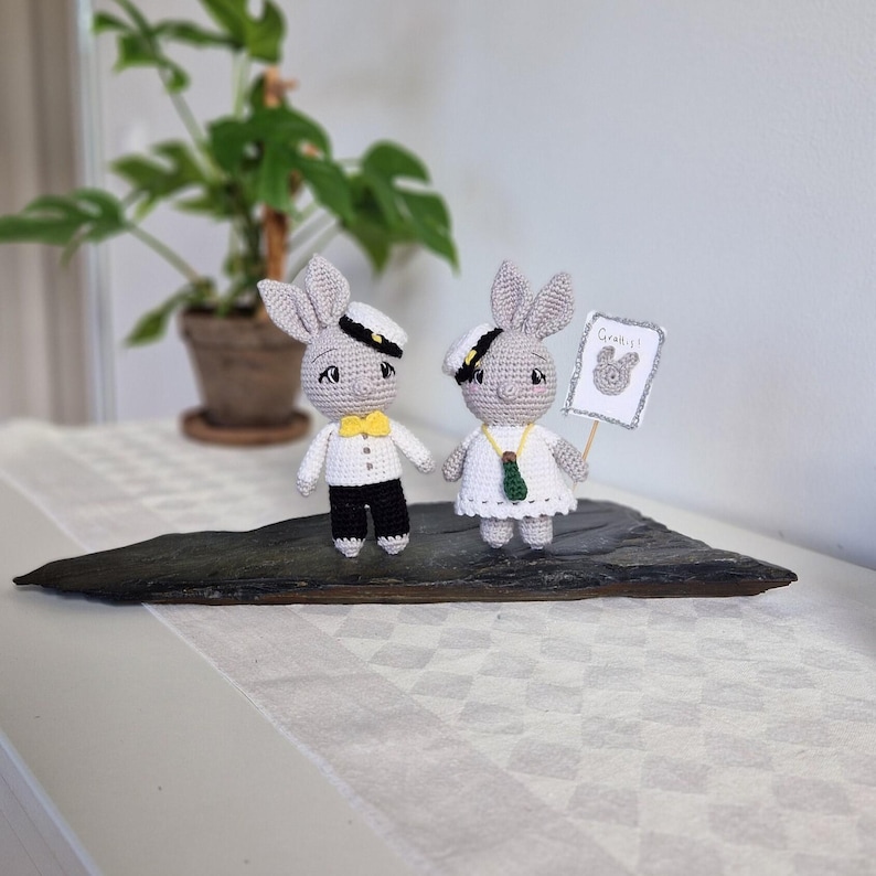 May include: Two crocheted bunny figurines dressed in sailor outfits. The bunny on the right is holding a sign that says "Grattis!" in white lettering on a light blue background.