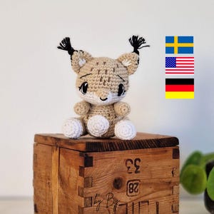 Op de afbeelding: Een gehaakt lynx knuffeldier met zwarte oren en witte pootjes, zittend op een houten doos met de tekst "by Bg" en de nummers "28" en "33".