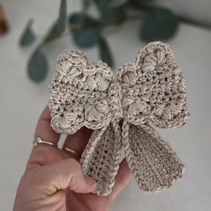 May include: A beige crocheted bow with a textured design. The bow features a detailed stitch pattern and two long, flowing tails. The handmade bow is a decorative accessory.