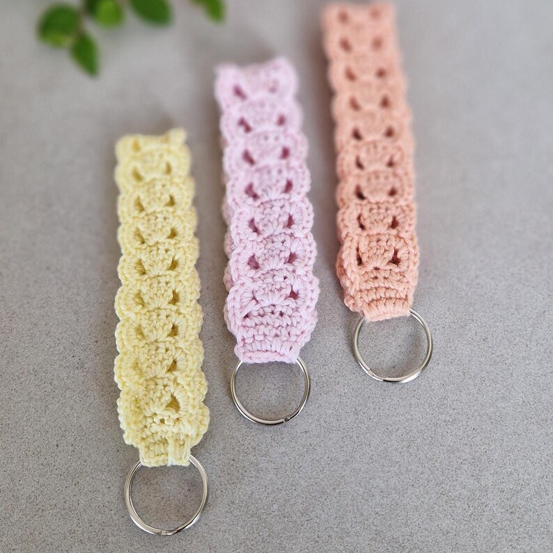 Op de afbeelding: Drie handgemaakte gehaakte sleutelhangers in pastelkleuren: geel, roze en perzik. Elke sleutelhanger heeft een decoratief openwerkpatroon en een zilverkleurige metalen ring om sleutels aan te bevestigen. De sleutelhangers worden getoond op een neutrale grijze ondergrond.