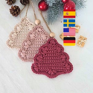May include: Three handmade crochet Christmas tree ornaments in shades of beige, pink, and burgundy. Each tree has a scalloped edge and a wooden bead for hanging. The ornaments are displayed on a white surface with festive decorations.