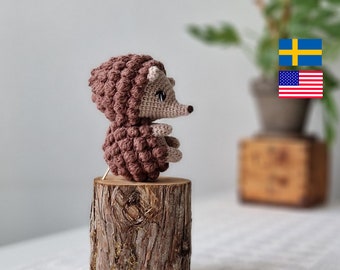 May include: A brown crocheted hedgehog sitting on a tree stump. The hedgehog has a light brown face with black eyes and a small pink nose.