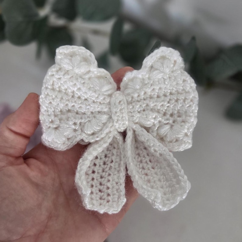 May include: A handmade, white crochet bow with a detailed floral design. The bow is held in a hand, showcasing the intricate stitching and delicate texture. The bow's shape is symmetrical, with two loops and a central knot.