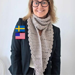 Könnte beinhalten: Ein beigefarbener, dreieckiger Häkelschal mit einem komplizierten Lochmuster und einem gewellten Rand mit kleinen Büschelmaschen-Details. Der Schal ist um den Hals über einem schwarzen Blazer und einem weißen Oberteil drapiert, mit schwedischen und amerikanischen Flaggensymbolen.