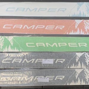 May include: Five white vinyl decals with a mountain and tree design. Each decal has the word "CAMPER" printed in black, white, or green.