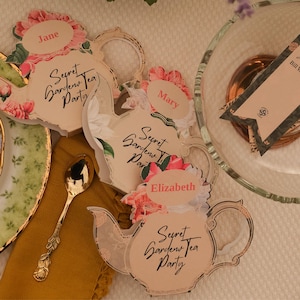 Può includere: Tre segnaposto di carta a forma di teiera con decorazioni floreali e la scritta Secret Garden Tea Party, con i nomi Jane, Mary ed Elizabeth. È visibile un cucchiaio in metallo dorato con il manico decorato a motivi di foglie.