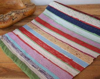 May include: A colorful, striped rag rug with a green border. The rug features a variety of colors, including red, blue, pink, and beige. The rug is made of woven fabric and is displayed on a wooden surface.