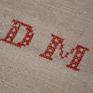 以下が含まれることがあります： 白いリネン生地のクローズアップ。赤い糸で「DM」の文字が刺繍されています。