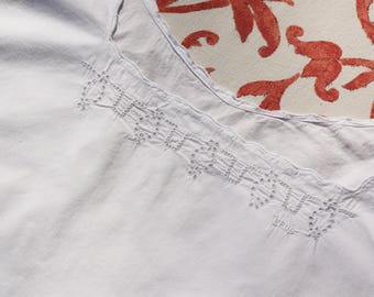 Puede incluir: Primer plano de una blusa blanca de estilo vintage con intrincados detalles bordados a lo largo del escote. La tela parece suave y ligeramente texturizada. El fondo presenta un estampado floral en tonos rojos y crema.