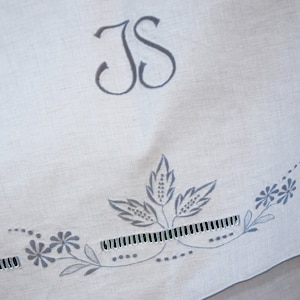 May include: White linen fabric with a gray embroidered monogram "JS" and a gray floral design along the bottom edge.