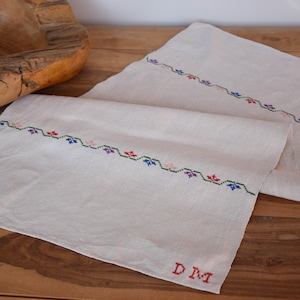 Könnte beinhalten: Ein weißer Tischläufer aus Leinen mit gestickten Blumendesigns in Rot, Blau, Grün und Rosa. Der Läufer trägt die Initialen "D.M." in Rot gestickt in der unteren rechten Ecke.