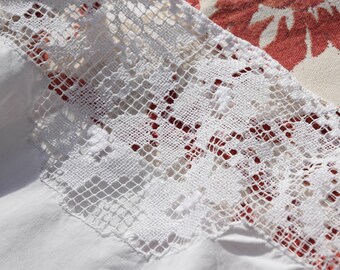 Puede incluir: Primer plano de un ribete de encaje blanco sobre una tela blanca. El encaje tiene un diseño calado delicado. El fondo presenta un estampado floral en rojo y crema.
