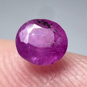 Può includere: Una gemma ovale di colore rosa vibrante con taglio sfaccettato. La gemma presenta inclusioni nere visibili all'interno della pietra.