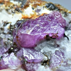 Puede incluir: Primer plano de una gema en bruto, sin cortar, con un vibrante tono rosa. La estructura cristalina es visible, sobre un fondo de minerales blancos y beige. La textura rugosa y la formación natural son prominentes.