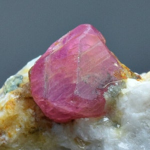 Op de afbeelding: Een levendig roze robijnkristal met een veelzijdig, gefacetteerd oppervlak. Het kristal is ingebed in een matrix van wit en bruin gesteente.