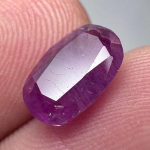 Pode incluir: Uma gema púrpura com forma oval facetada. A gema tem um tom púrpura claro e é translúcida.