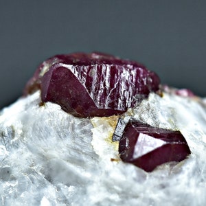 Può includere: Due cristalli di rubino rosso scuro su una matrice bianca. I cristalli sono affilati e hanno una lucentezza vitrea.