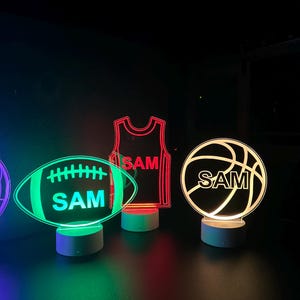 May include: Three illuminated acrylic night lights shaped like a basketball, football, and basketball jersey. Each light has the name "SAM" inscribed on it. The basketball is white with black lines, the football is green with white lines, and the jersey is red.