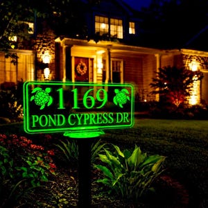 May include: A rectangular, glowing green address sign mounted on a black stake. The sign features the number 1169, the text POND CYPRESS DR, and two sea turtle graphics, one on each side of the house number, all illuminated in bright green light.