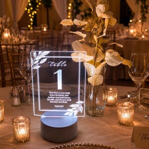 May include: A rectangular clear acrylic table number sign labeled "Table 1" with white botanical line art and the text "Therefore a man shall leave his father and mother and hold fast to his wife, and the two shall become one flesh. Mark 10:7" illuminated on a round base.