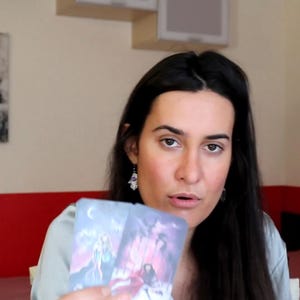 May include: A person holding two tarot cards with colorful illustrations. The cards feature images of figures and celestial bodies. The person is wearing a light blue shirt and has long, dark hair. A painting hangs on the wall behind them.