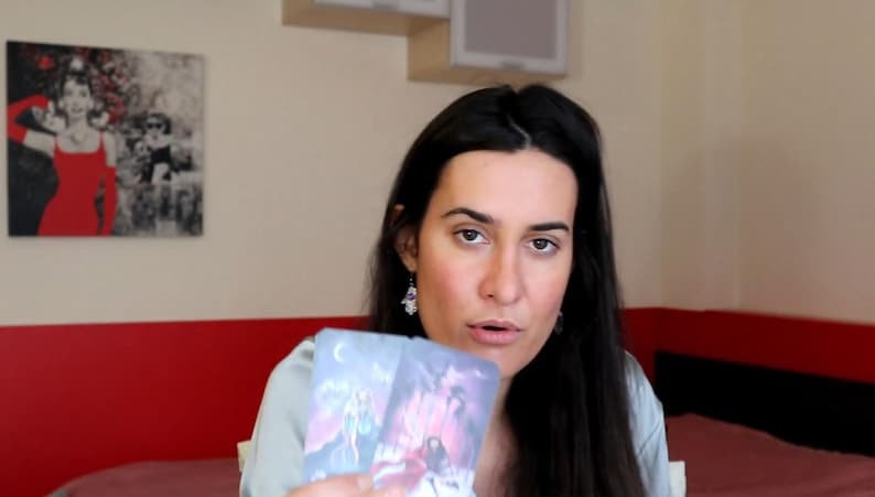 May include: A person holding two tarot cards with colorful illustrations. The cards feature images of figures and celestial bodies. The person is wearing a light blue shirt and has long, dark hair. A painting hangs on the wall behind them.