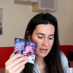 May include: Two tarot cards are held up, displaying colorful illustrations of figures and celestial elements. The cards feature a dark blue and purple color scheme with white and red accents. The image is of a person holding the cards.