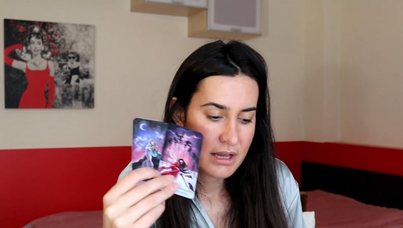 May include: Two tarot cards are held up, displaying colorful illustrations of figures and celestial elements. The cards feature a dark blue and purple color scheme with white and red accents. The image is of a person holding the cards.
