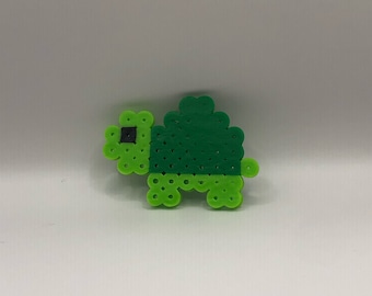 Op de afbeelding: Een groene en zwarte kralen schildpad, gemaakt met kleine ronde kralen. De schildpad heeft een groene schild en een zwart oog.
