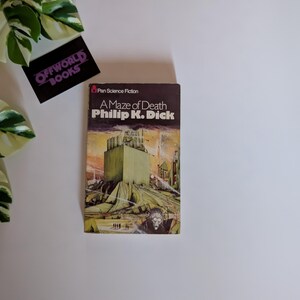 May include: A vintage paperback book titled "A Maze of Death" by Philip K. Dick. The cover features a futuristic illustration in shades of brown, green, and yellow. The book is a Pan Science Fiction edition. A black card with "OFFWORLD BOOKS" is also visible.