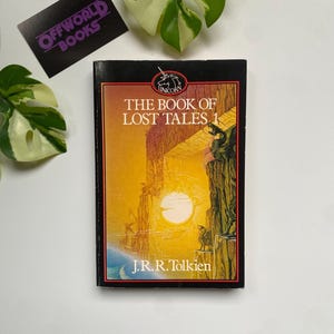 K&ouml;nnte beinhalten: Ein gebundenes Buch mit dem Titel "The Book of Lost Tales 1" von J.R.R. Tolkien. Das Cover zeigt eine Sonnenuntergangsszene mit Figuren auf einer Klippe. Das Buch hat einen schwarzen Buchr&uuml;cken und liegt vor einem wei&szlig;en Hintergrund mit gr&uuml;nen Pflanzen.