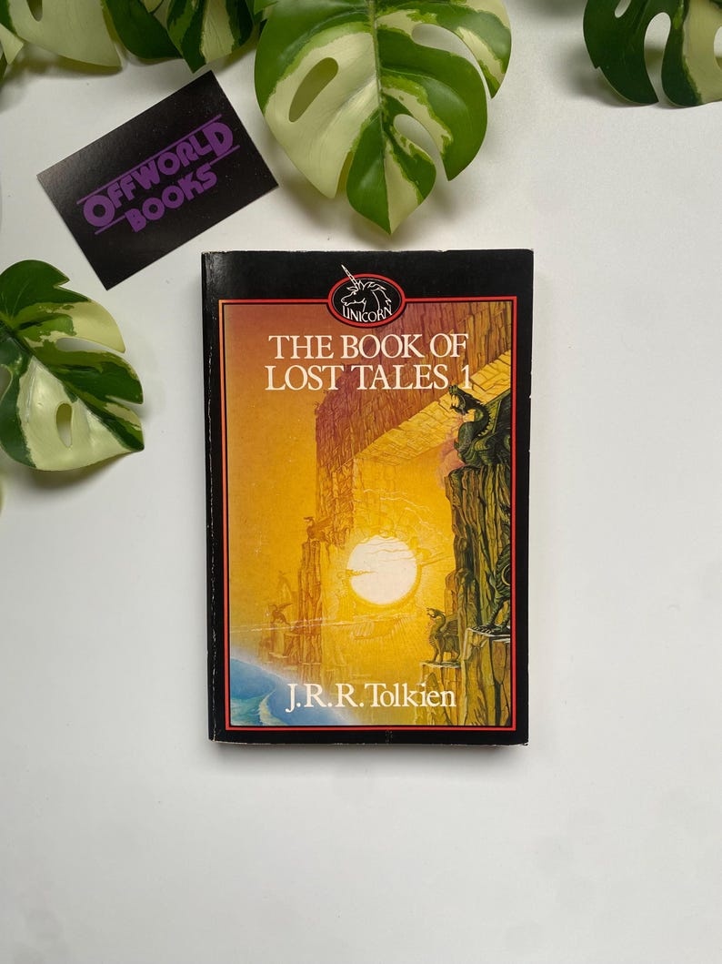 K&ouml;nnte beinhalten: Ein gebundenes Buch mit dem Titel "The Book of Lost Tales 1" von J.R.R. Tolkien. Das Cover zeigt eine Sonnenuntergangsszene mit Figuren auf einer Klippe. Das Buch hat einen schwarzen Buchr&uuml;cken und liegt vor einem wei&szlig;en Hintergrund mit gr&uuml;nen Pflanzen.