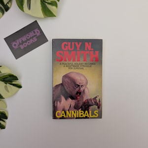 May include: A paperback book titled Cannibals by Guy N. Smith with a cover illustration of a pale, monstrous creature. The text reads: A peaceful holiday becomes a nightmare struggle for survival. A black card with the text Offworld Books is nearby.
