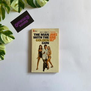 May include: A vintage paperback book titled "The Man with the Golden Gun" by Ian Fleming, featuring James Bond. The cover art depicts three figures. A black and purple "OFFWORLD BOOKS" sticker is in the upper left corner.