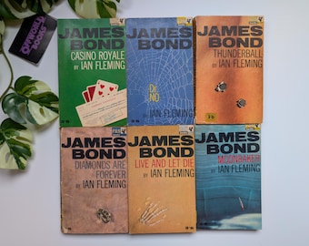 Könnte beinhalten: Eine Sammlung von sechs alten James Bond Taschenbüchern von Ian Fleming, darunter Casino Royale, Dr. No, Thunderball, Diamonds Are Forever, Live and Let Die und Moonraker, jeweils mit eigenem Cover-Design und dem Pan Books Logo.