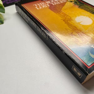 K&ouml;nnte beinhalten: Ein Hardcover-Buch mit dem Titel "The Book of Lost Tales 1" mit schwarzem Buchr&uuml;cken und einem farbenfrohen Cover mit einer Landschaftsszene mit Sonne. Das Cover des Buches hat einen Farbverlauf von Orange und Gelb.