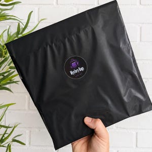 May include: A square black plastic shipping mailer bag featuring a circular sticker in the centre. The sticker has a purple and black graphic design with the text Mystery Bags printed in white letters across the middle of the circular label.