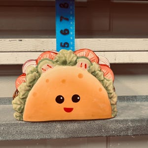 May include: A ceramic taco-shaped napkin holder with a smiling face, featuring painted lettuce, tomatoes, and onions. A blue ruler is positioned behind the item, showing a height measurement of approximately 5.5 inches from the base to the top.