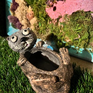May include: A whimsical, gray otter-shaped planter with large, expressive eyes and a wide open mouth. The planter is made of a textured, brown material and is set against a backdrop of green moss and a pink structure.