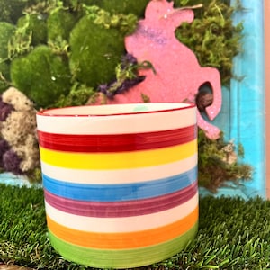 May include: A ceramic cylindrical mug with horizontal stripes in red, yellow, blue, purple, orange, and green. The mug has a white base and a red rim. The background includes a pink unicorn cutout and green moss.