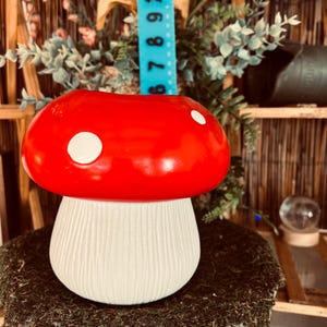 May include: A whimsical mushroom-shaped planter with a vibrant red cap and white polka dots. The stem is a textured, off-white color. This decorative piece adds a touch of fantasy to any garden or indoor space. The planter is approximately 6 inches tall.