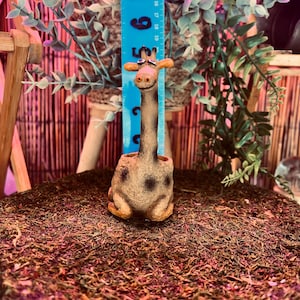 May include: A whimsical giraffe-shaped planter, featuring a long neck and a spotted body. The giraffe is tan with brown spots and has a smiling expression. A blue ruler is behind the giraffe. The planter is approximately 6 inches tall.
