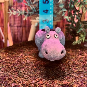 May include: A whimsical, hand-painted hippo planter in shades of pink and gray. The ceramic planter features a detailed hippo face with large eyes and a wide snout. A blue ruler with inch markings is visible in the background.