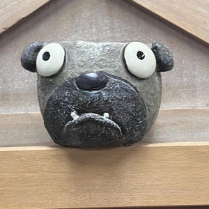 May include: A gray, pug-shaped planter with large white-rimmed eyes and a black nose. The planter has a textured surface and a wide, open mouth with two small teeth. The ears are rounded and stick out from the sides of the head. The planter is made of a durable material.