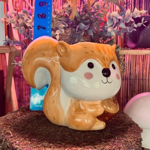 May include: A ceramic squirrel figurine with a cartoonish design. The squirrel has a white belly, brown and orange fur, and a large, curved tail. It has black eyes, pink cheeks, and is holding a nut. The figurine is on a bed of moss.