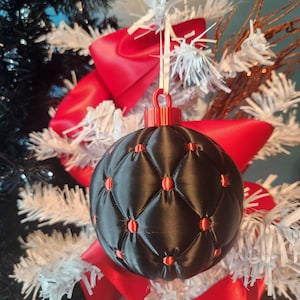 May include: A spherical Christmas ornament featuring a black quilted pattern with a matte finish. The intersection points of the diamond-shaped quilting are accented with small, glossy red circular studs. A red cap sits at the top.