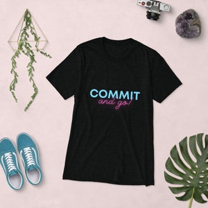 Könnte beinhalten: Schwarzes T-Shirt mit dem Schriftzug "COMMIT and go!" in Blau und Pink. Das Bild zeigt blaue Turnschuhe, eine Kamera und eine dekorative Pflanze auf einer rosa Oberfläche.