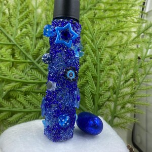 Puede incluir: Una botella de vidrio azul con tapa negra, decorada con cuentas azules y blancas, estrellas y amuletos de ojo turco. La botella está sobre una superficie blanca junto a un huevo azul con purpurina.
