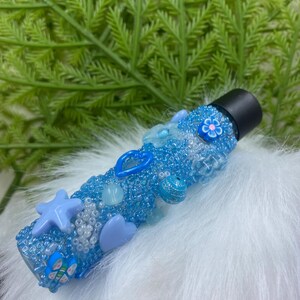 Puede incluir: Un frasco de vidrio azul con rodillo y tapa negra, decorado con cuentas azules y blancas, estrellas, corazones y flores.