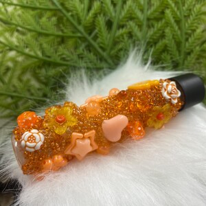 Puede incluir: Un tapón de botella decorativo de vidrio naranja y transparente, adornado con varios adornos. Estos incluyen cuentas naranjas, formas de estrella y corazón, pequeñas flores y una rosa blanca. El tapón tiene una parte superior negra y está sobre un fondo blanco y esponjoso.